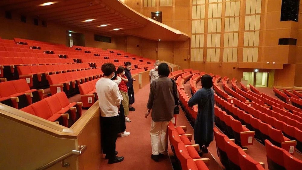 Project members tour Omuta Cultural Hall in Omuta City, Fukuoka Prefecture, a performance-hall-like space with rows of red seats facing the stage.