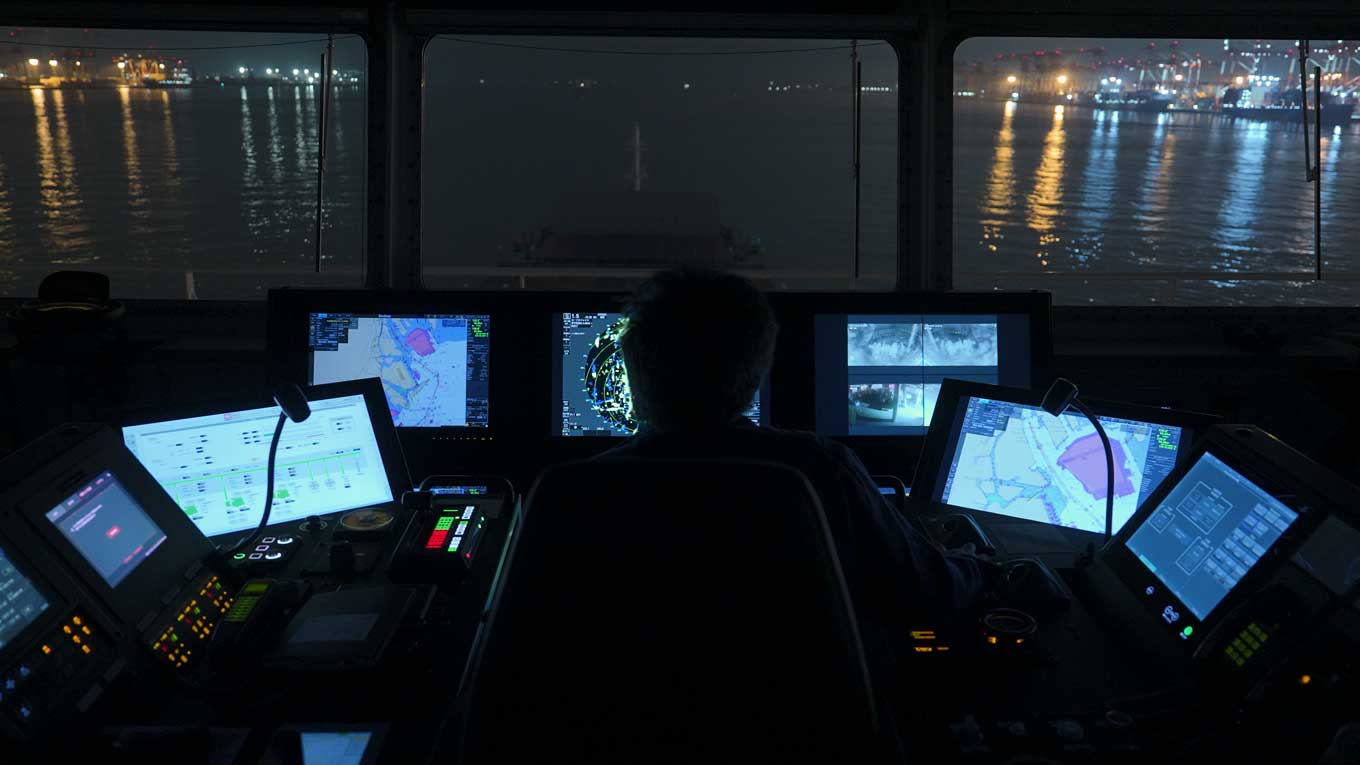 Photo: Nighttime view from GENBU’s bridge, where a crew member monitors multiple digital navigation screens as port lights reflect on the water.