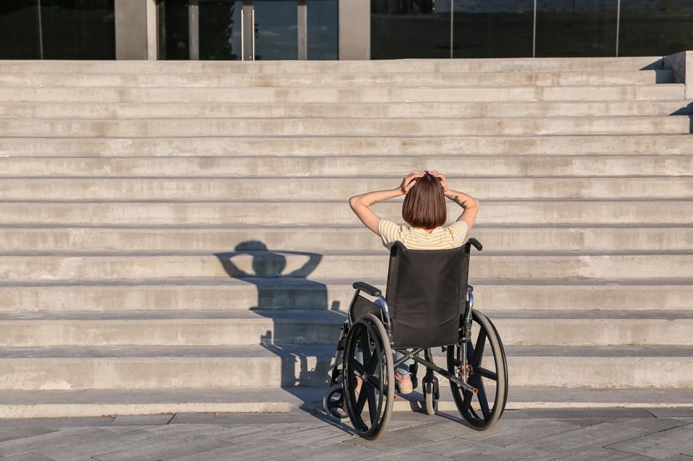 A wheelchair user holds her head in her hands facing a flight of stairs