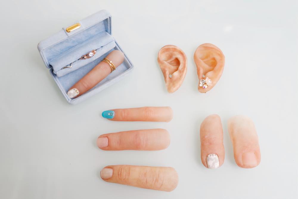 Lineup of epitheses: Three fingers (one with nail art), two toes (one with nail art), two ears (one with a pierced earring) and one jewelry case containing rings and a finger epithesis
