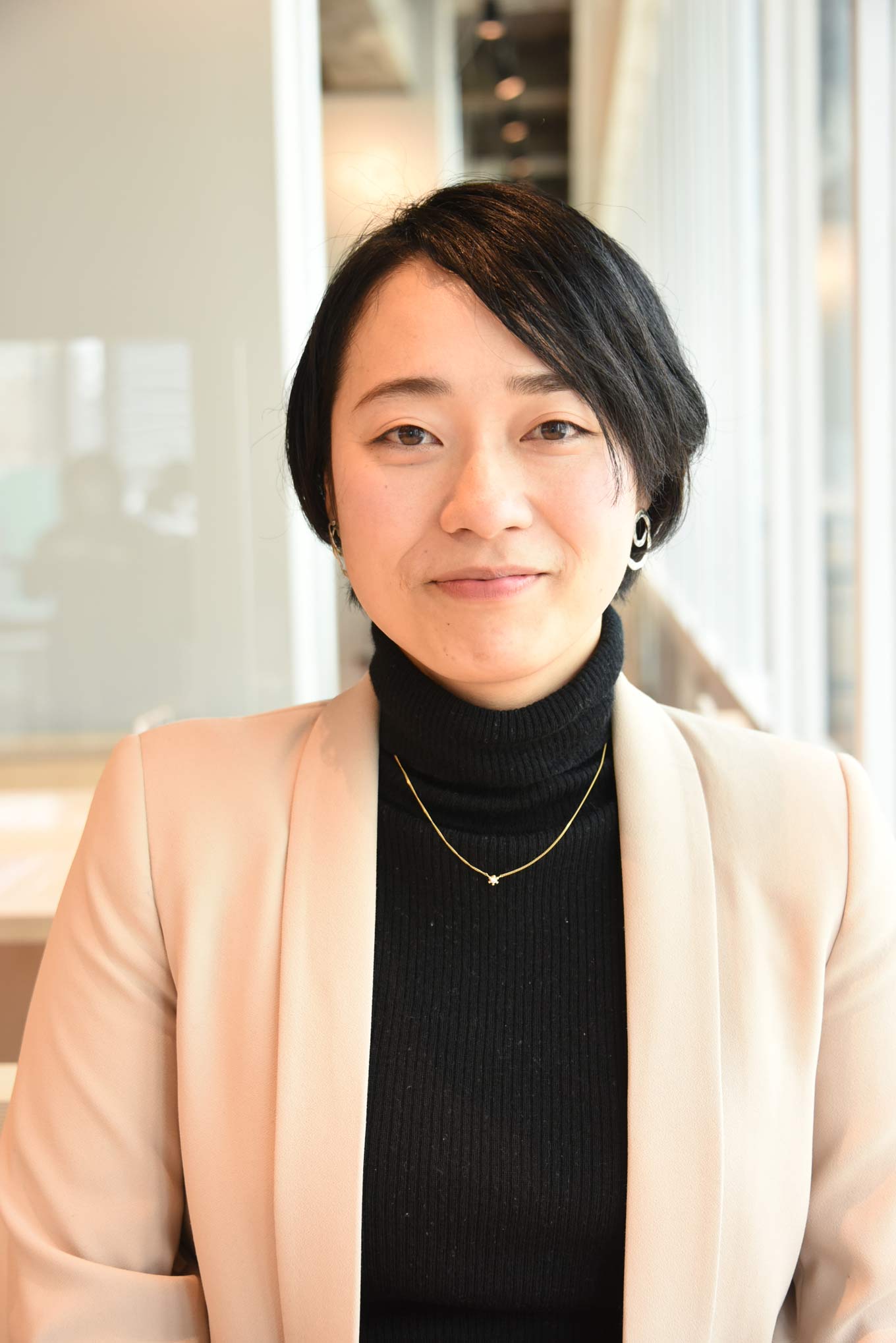Photo: Ikumi Kawamata smiles at the camera while standing indoors in a black turtleneck and dark blazer.