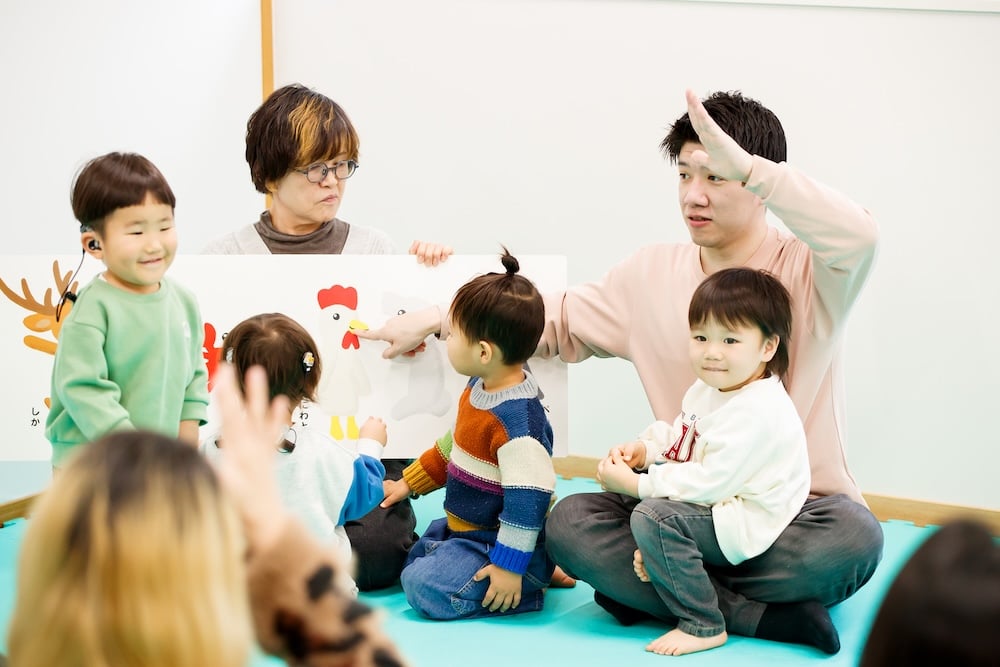 Children gather around Yutaka Kubosawa as he explains a drawing in sign language; one child is even sitting on his lap
