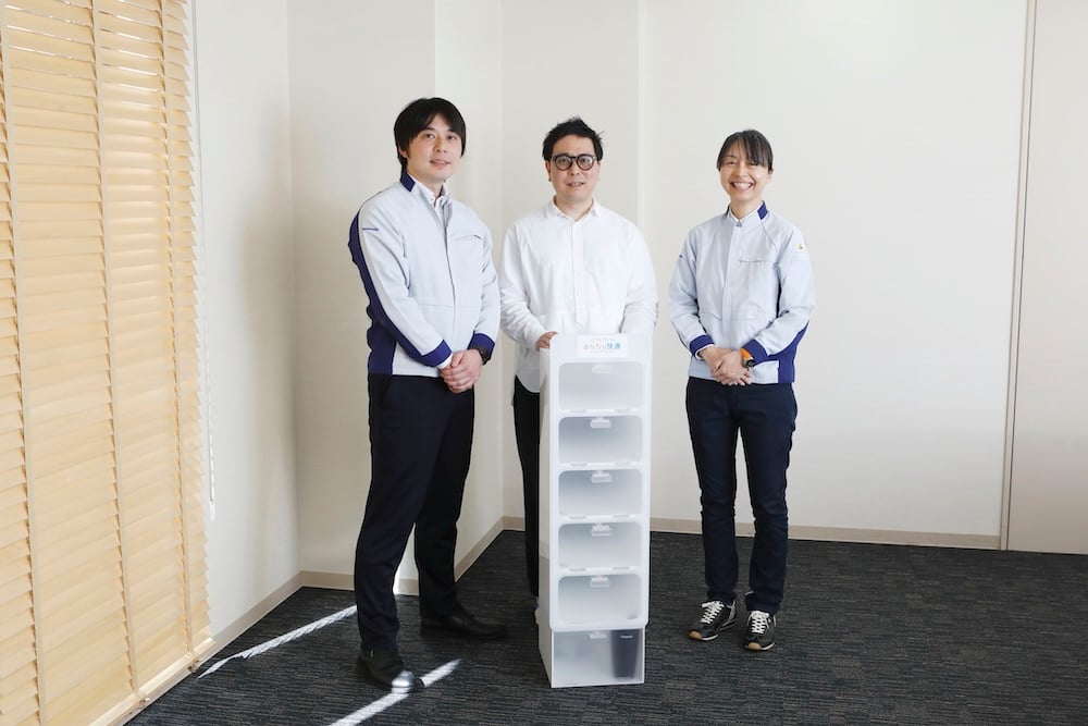 In a corner of a room, Jumpei Oe, Takashi Watanabe, and Sawako Nakagawa stand side by side, with a white shoe cabinet in front of them.