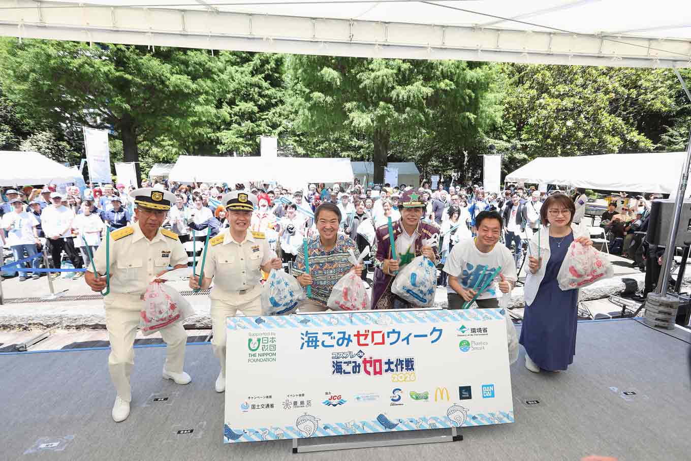 Photo: With a large crowd gathered behind them, participants and event representatives stand onstage holding litter pickers and bags of collected rubbish at the kickoff event for UMIGOMI Zero Week in Tokyo